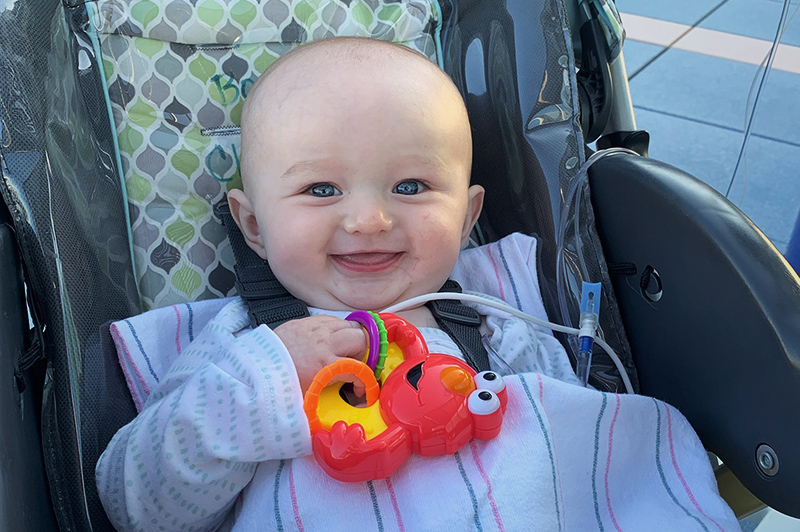 Smiling baby sitting in car seat is covered by striped blanket and holds Elmo rattle