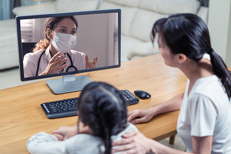 Clinician wearing mask is shown on monitor; young girl and adult woman look at clinician during virtual visit