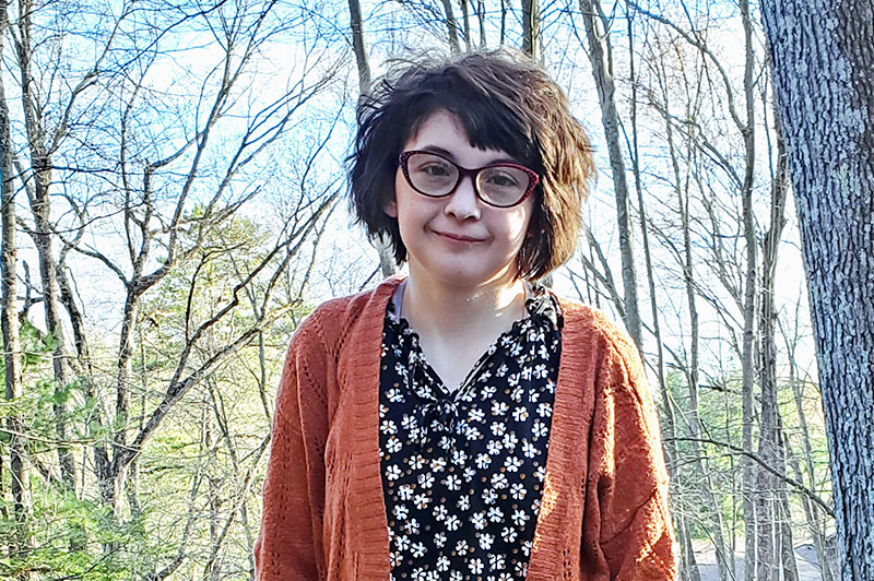 Young girl wearing glasses and a cardigan sweater stands in wooded area